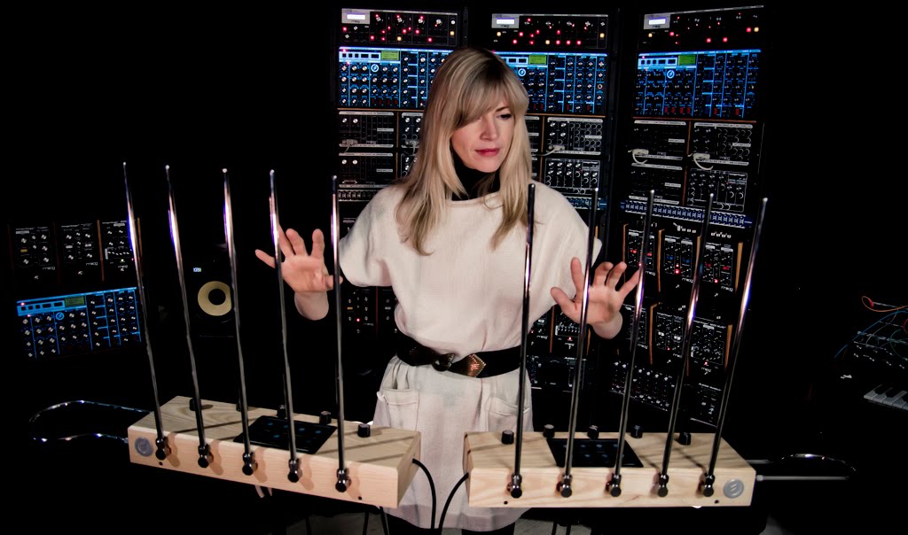 Mulher loira com roupa futurista tocando dois theremins com 5 antenas cada em uma sala com vários equipamentos elétricos (sintetizadores) ao fundo.