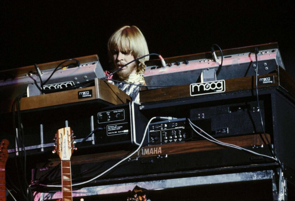 Rick Wackerman do Yes em uma apresentação ao vivo, com dois minimoogs. A foto mostra braços de um violão e uma guitarra e a parte de trás de equipamentos de som. Rick Wackerman está tocando um minimoog.